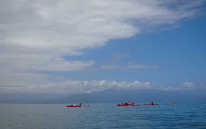 A line of red kayaks are paddled across the calm blue ocean. There are mountains in the distance behind them.